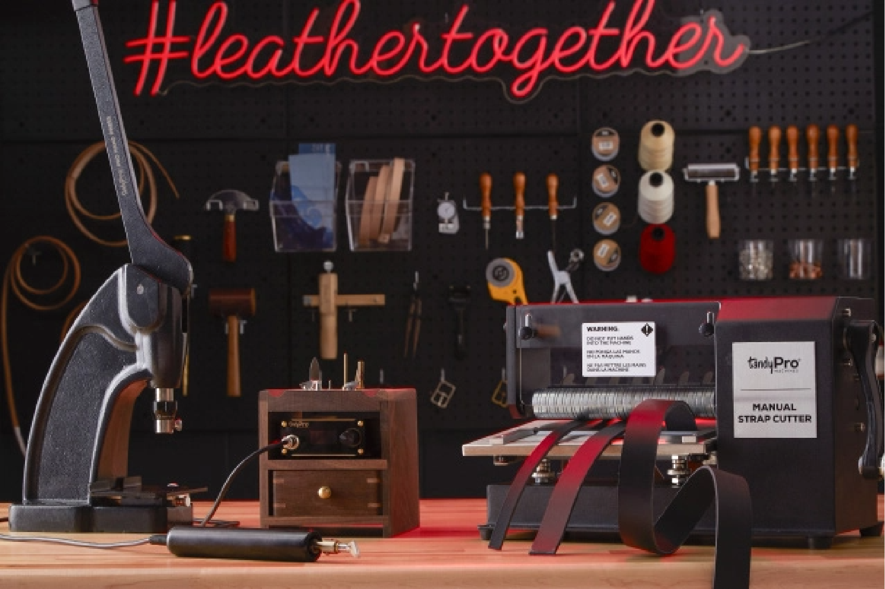 Leather tools, a hand press and a TandyPro manual strap cutter on a workbench beneath a #leathertogether neon sign